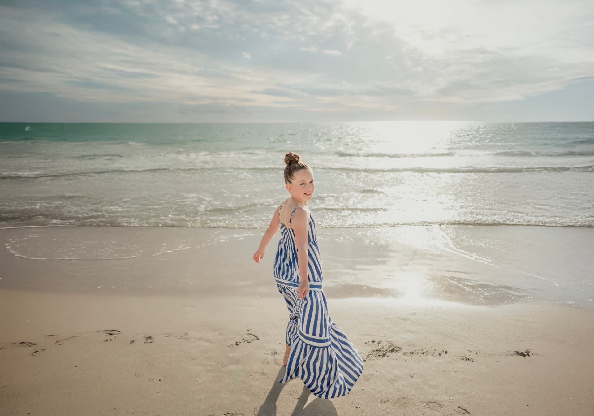 Olivia glancing back with a smile as she walks along the wet sand in a striped dress