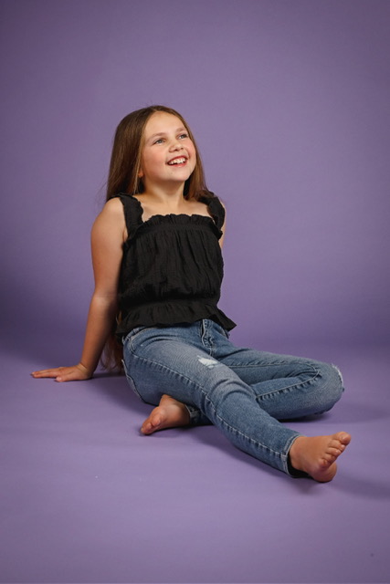 Olivia sitting on a purple studio backdrop in a black top and jeans, smiling up at the light