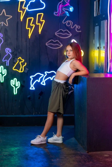 Olivia posing beside colorful neon artwork in a white top and green shorts