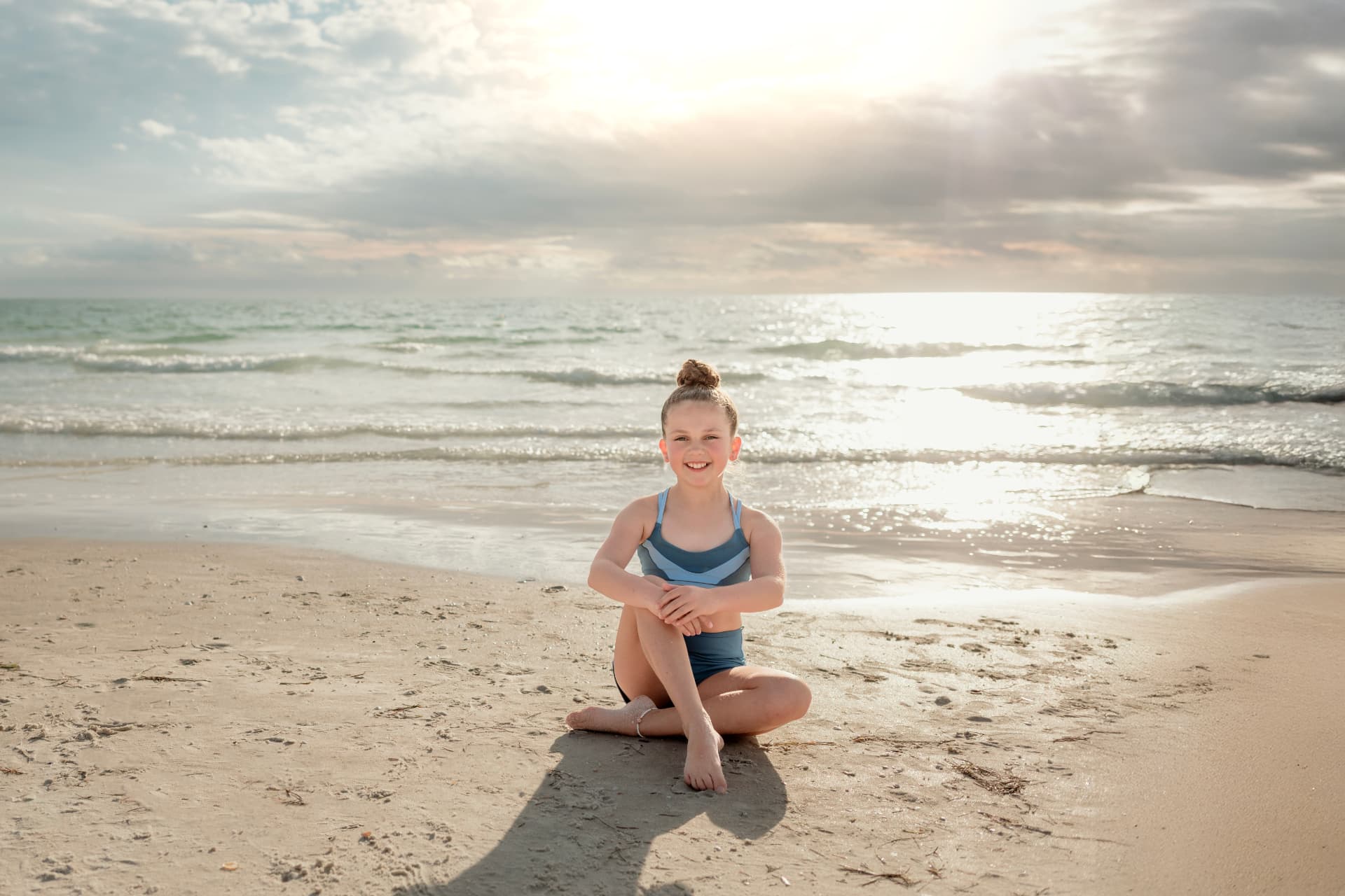 Olivia sitting cross-legged on the beach in a blue dance outfit, backlit by the sun
